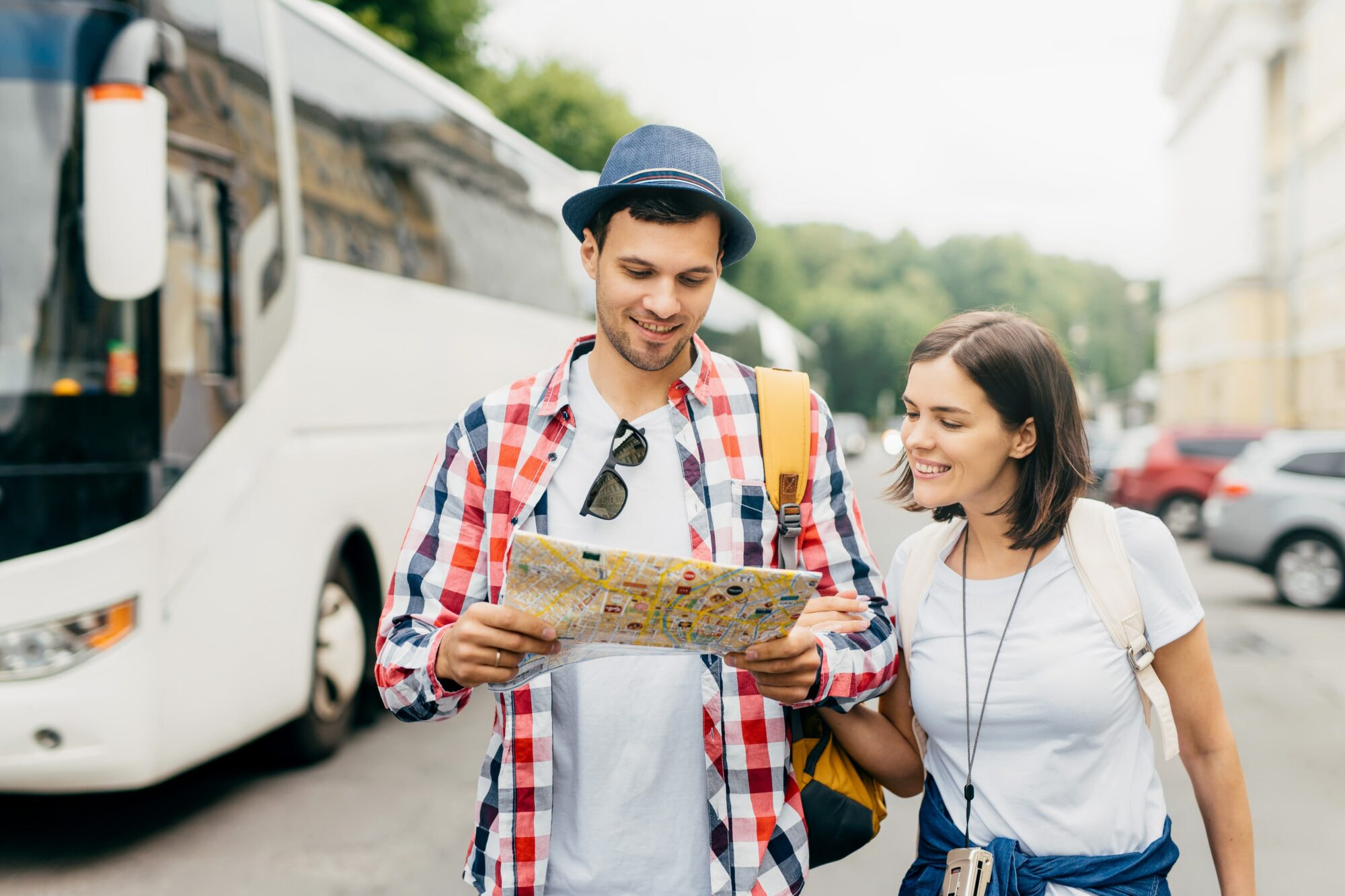 Cheerful guy in hat and shirt, holding city map, looking with smile in it, standing near his female friend, walking in city, exploring new entertainments. Male and female tourists with city guide.