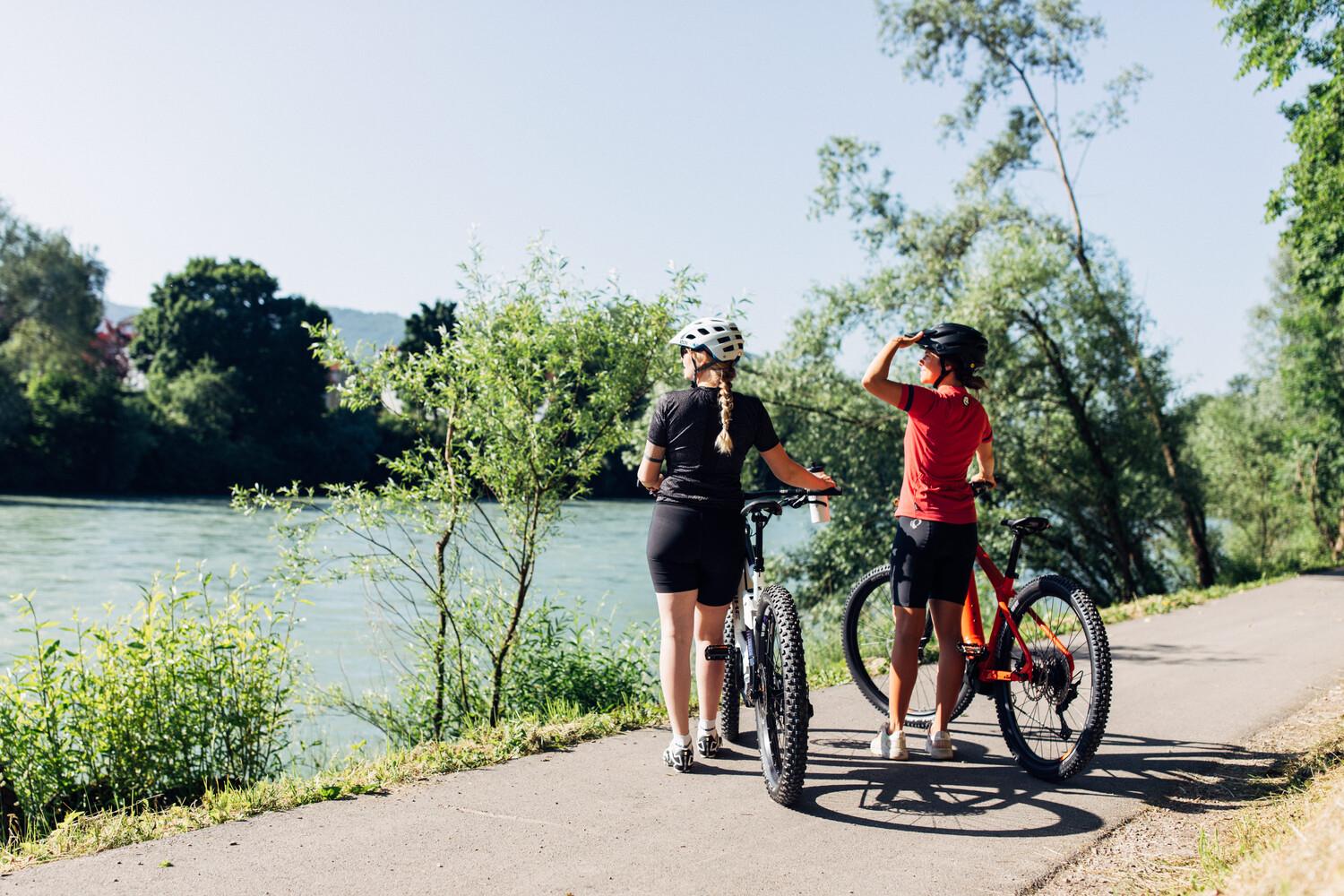 Zwei Frauen stehen mit ihren Fahrrädern in der Hand auf einem schmalen Weg mit Blick auf einen Fluss