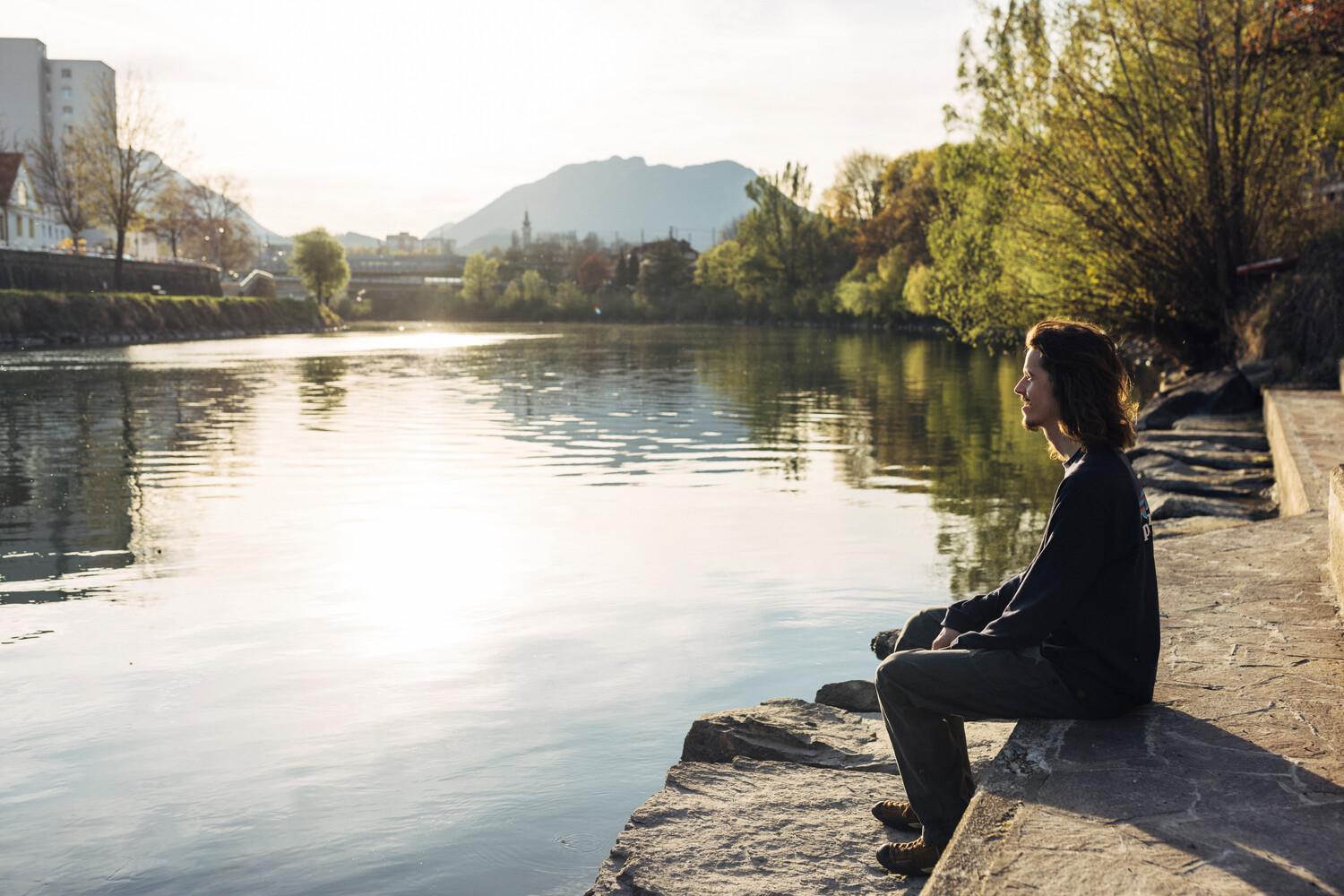 Ein junger Mann sitzt bei gutem Wetter am Rande eines Flusses und genießt die Sonne