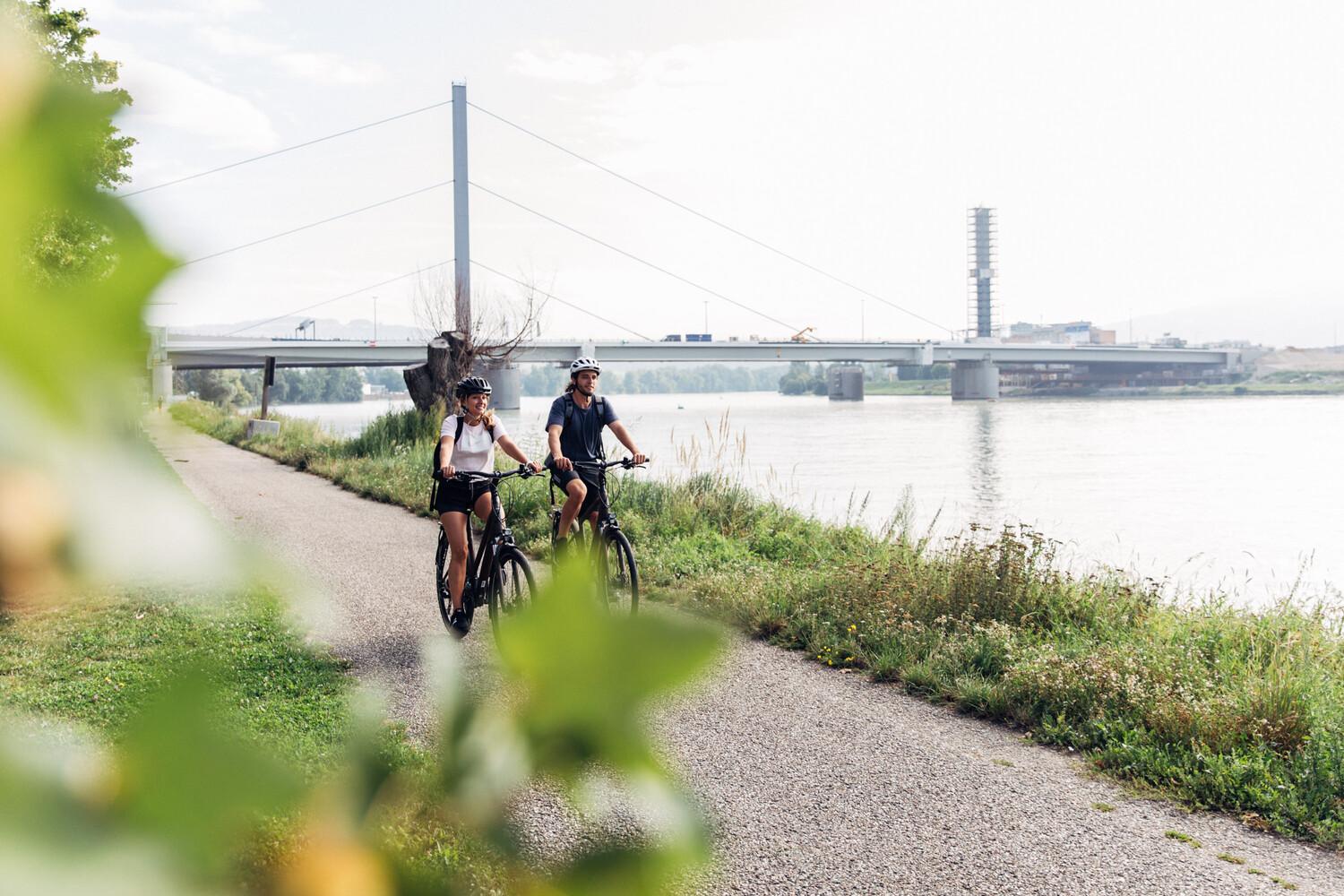 Zwei junge Menschen fahren nebeneinander auf Fahrrädern an einem Fluss entlang