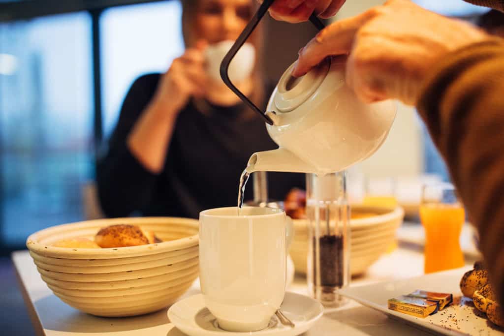 Person gießt sich Tee in eine Tasse ein an gefülltem Frühstückstisch mit einer Frau gegenübersitzend