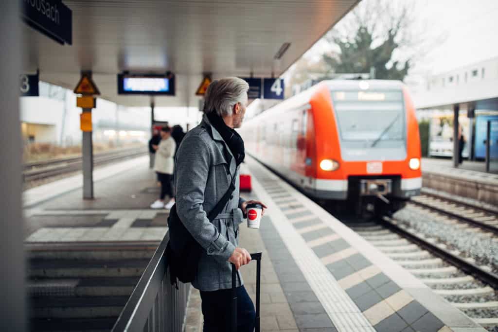 Mann mit Koffer und Kaffee in der Hand wartet an einem Bahngleis auf die gerade einfahrende S-Bahn