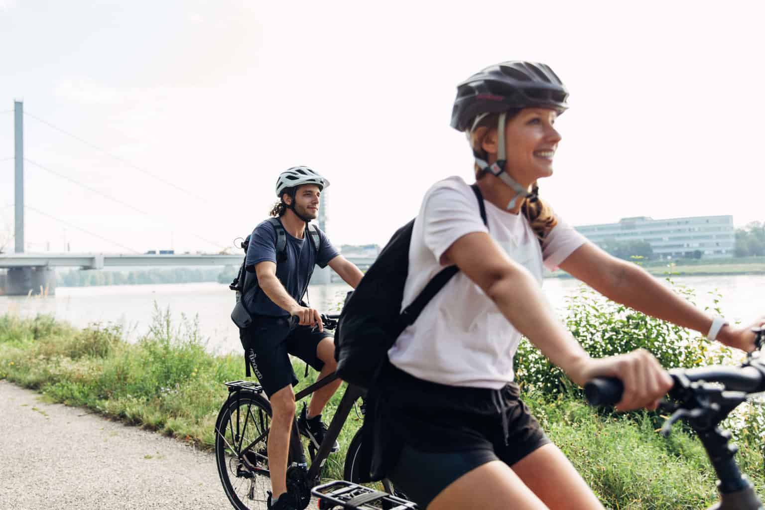 Zwei Personen fahren mit Helmen auf ihren Fahrrädern an einem Fluss entlang