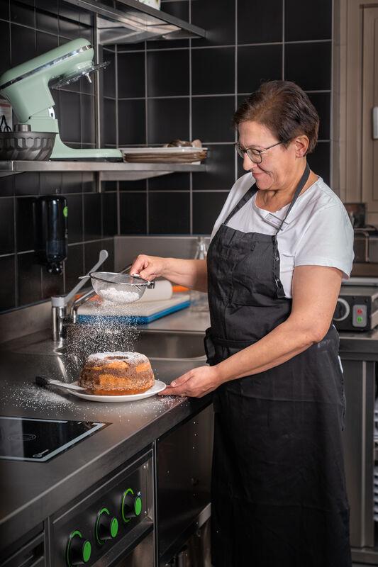 Eine Frau steht in einer Küche mit Schürze und streut einen Kuchen mit Puderzucker ein